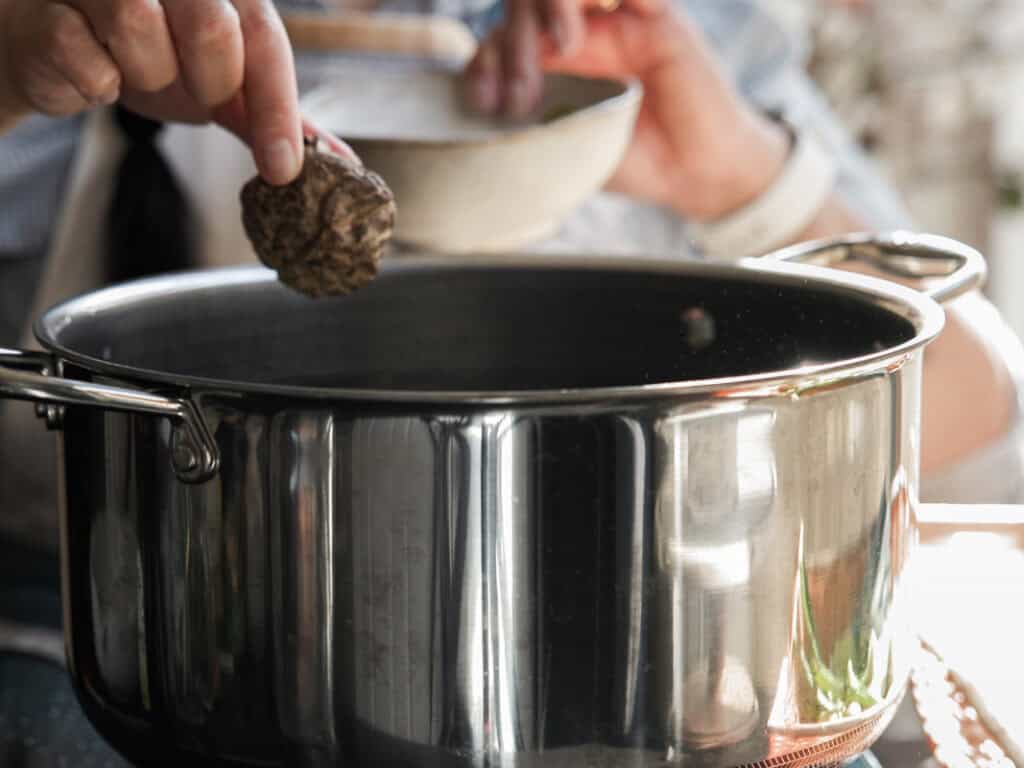 A person holds a mushroom and a bowl over a shiny metal pot on a stove, suggesting cooking or preparing a meal. The focus is on the mushroom and pot, with soft natural light illuminating the scene.